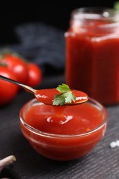 Photo of Taking tasty ketchup with spoon from bowl at black wooden table, closeup. Tomato sauce