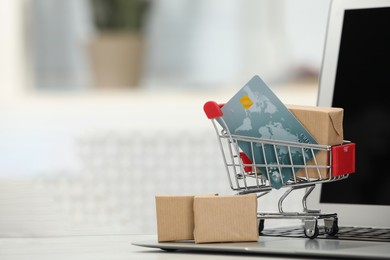 Online payment concept. Small shopping cart with bank card, boxes and laptop on table, space for text