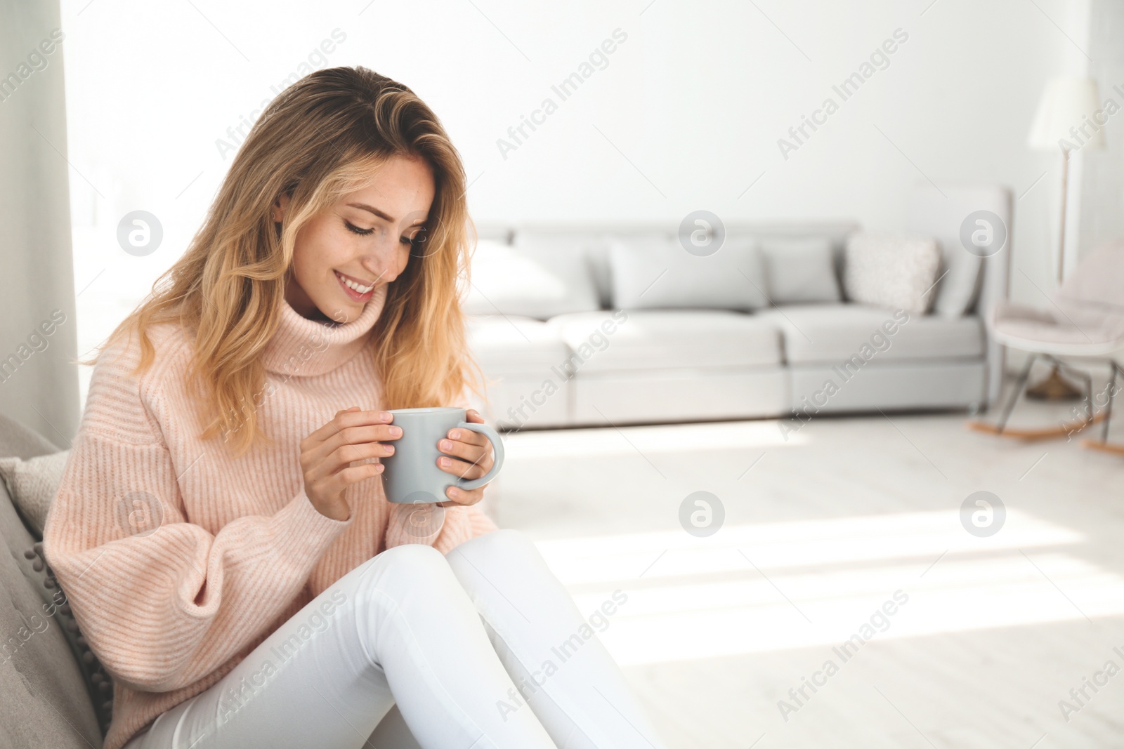 Photo of Beautiful young woman with cup of hot drink wearing warm pink sweater at home. Space for text