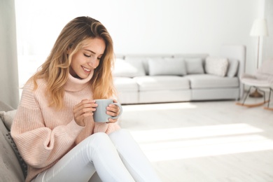 Beautiful young woman with cup of hot drink wearing warm pink sweater at home. Space for text