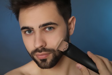 Handsome young man shaving with electric trimmer on blue background, closeup