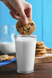 Photo of Woman dipping delicious chocolate chip cookie into glass of milk at wooden table, closeup