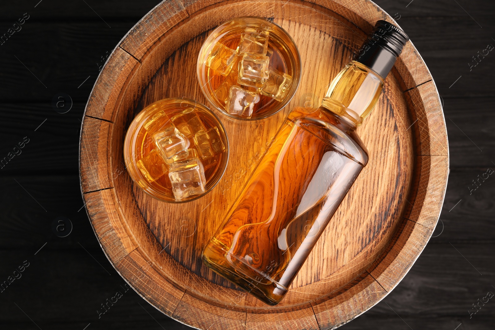 Photo of Whiskey with ice cubes in glasses, bottle and barrel on black wooden table, top view