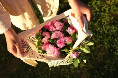 Woman holding mesh bag with beautiful tea roses outdoors, top view