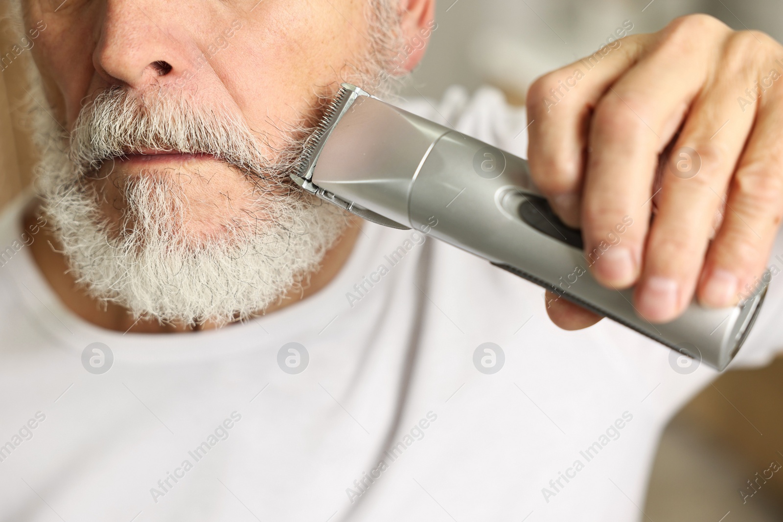 Photo of Man trimming beard with electric trimmer indoors, closeup