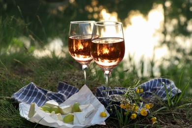 Glasses of delicious rose wine, cheese and grapes on picnic blanket near lake