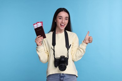 Happy woman with passport, tickets and camera showing thumb up on light blue background