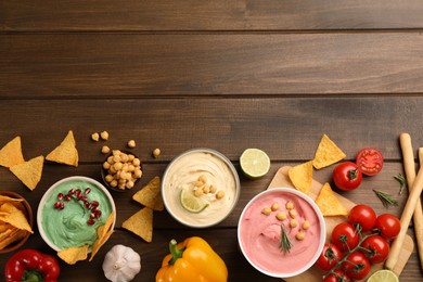 Photo of Different kinds of tasty hummus, nachos and ingredients on wooden table, flat lay. Space for text