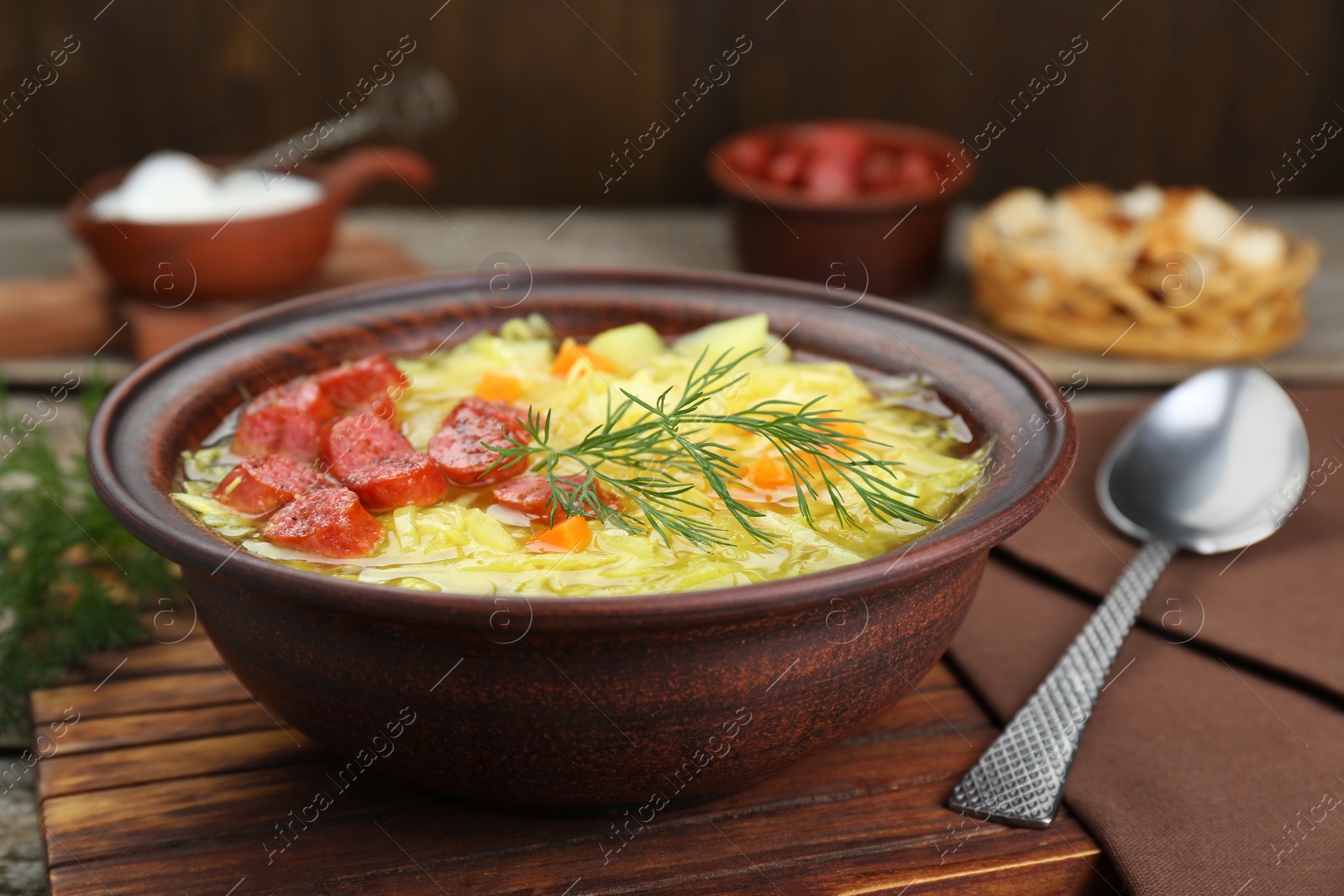 Photo of Bowl of delicious sauerkraut soup with smoked sausages and dill on wooden board