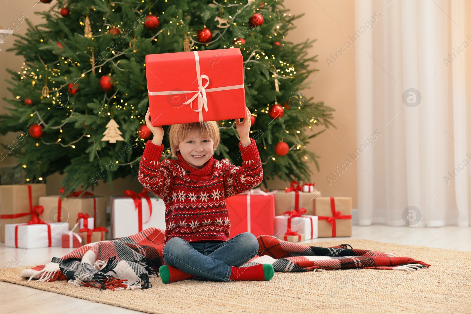 Photo of Little child with gift box on floor near Christmas tree at home