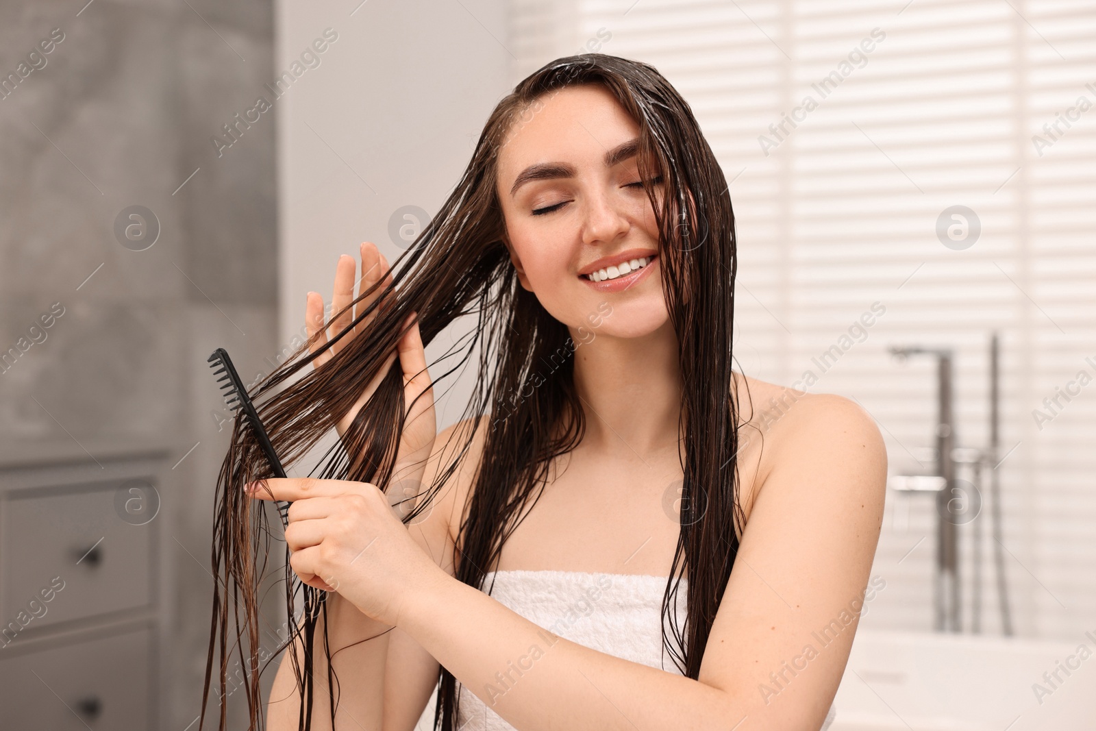 Photo of Young woman brushing hair after applying mask in bathroom