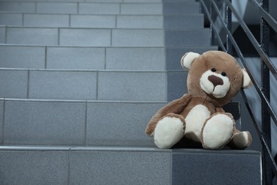 Photo of Lonely teddy bear on staircase near metal railing indoors, space for text