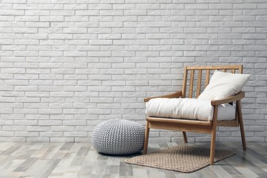 Stylish knitted pouf and comfortable armchair near white brick wall indoors, space for text. Interior design