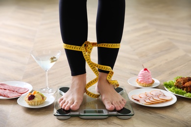Photo of Food, alcohol left after holidays and woman with measuring tape standing on scales indoors, closeup. Overweight problem