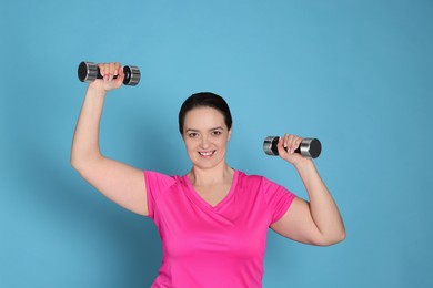 Happy overweight woman doing exercise with dumbbells on light blue background
