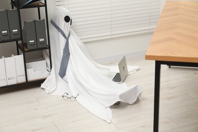 Photo of Overworked ghost. Man in white sheet with laptop on floor in office