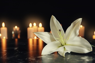 Photo of White lily and blurred burning candles on table in darkness, space for text. Funeral symbol