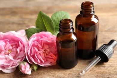 Photo of Bottles of rose essential oil, pipette and flowers on wooden table