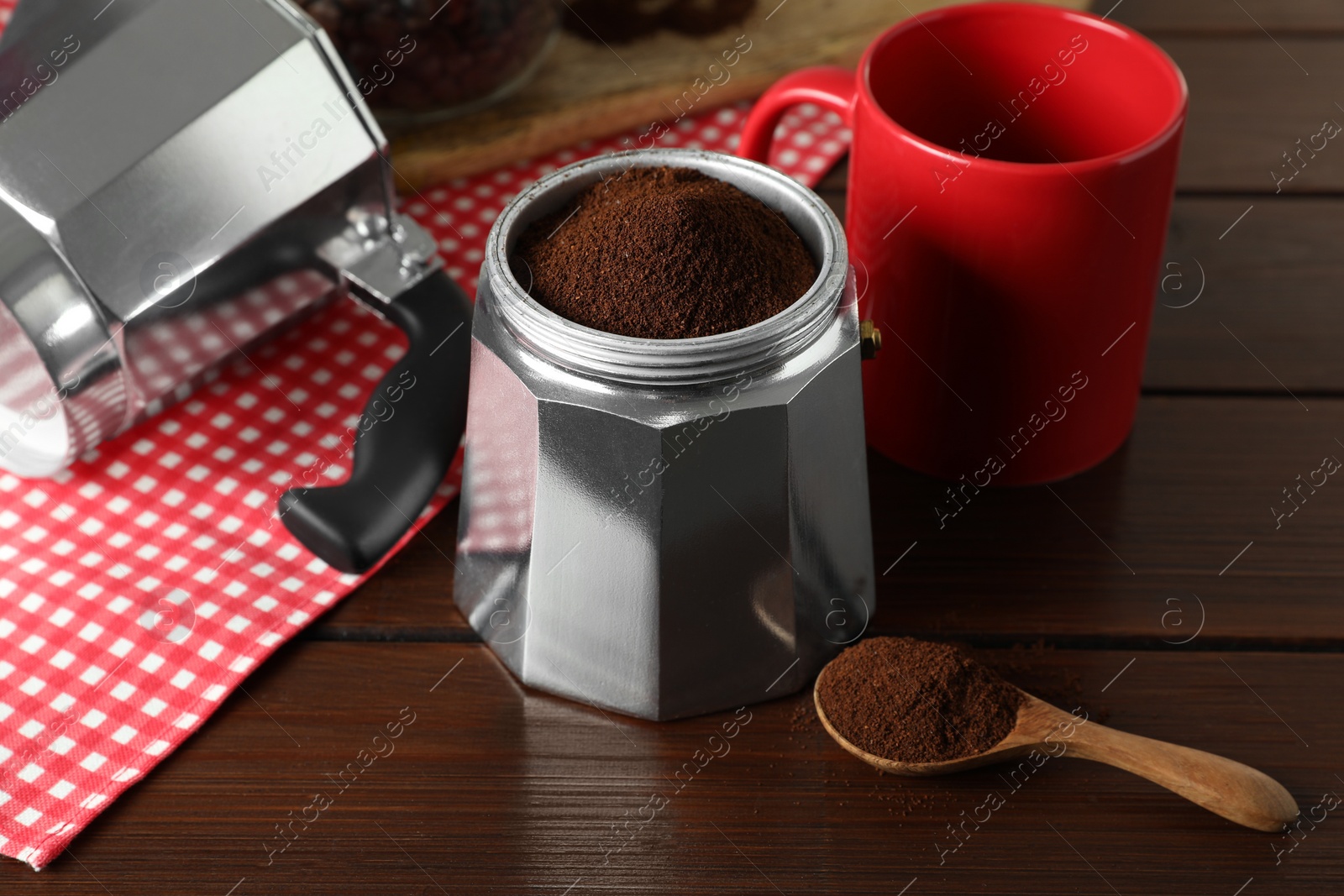 Photo of Moka pot with ground coffee, spoon and red cup on wooden table