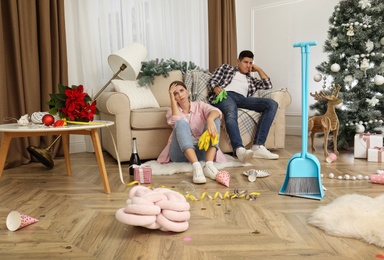 Photo of Tired couple sitting in messy room while cleaning after New Year party