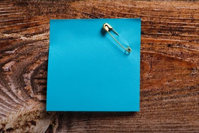Light blue paper note attached with safety pin on wooden table, top view. Space for text