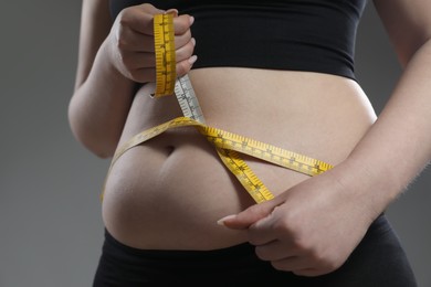 Woman measuring belly with tape on grey background, closeup. Overweight problem