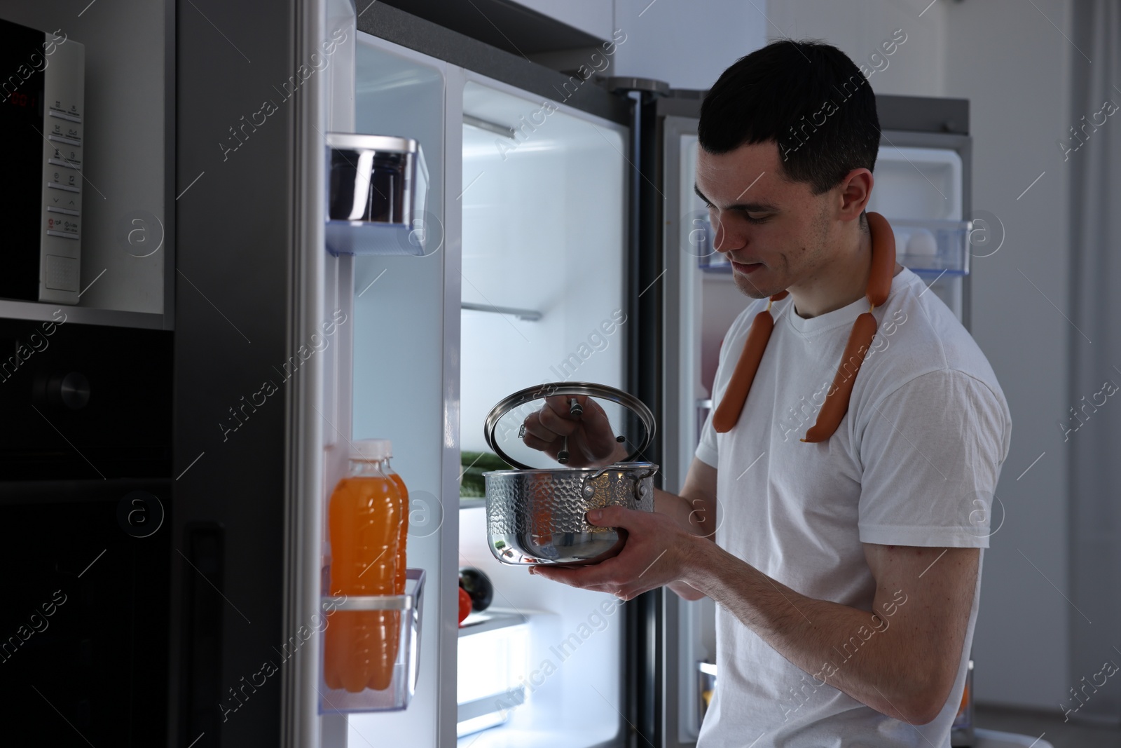 Photo of Man with sausages and pot near refrigerator in kitchen at night