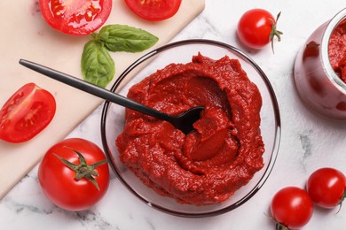 Photo of Glass bowl of tasty tomato paste with spoon and ingredients on white marble table, flat lay