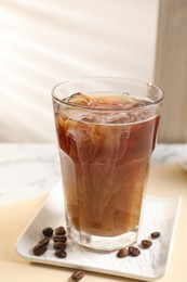 Refreshing iced coffee with milk in glass and beans on beige table