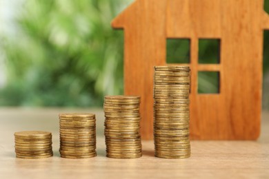 Mortgage concept. Stacks of coins and house model on wooden table against blurred green background, closeup
