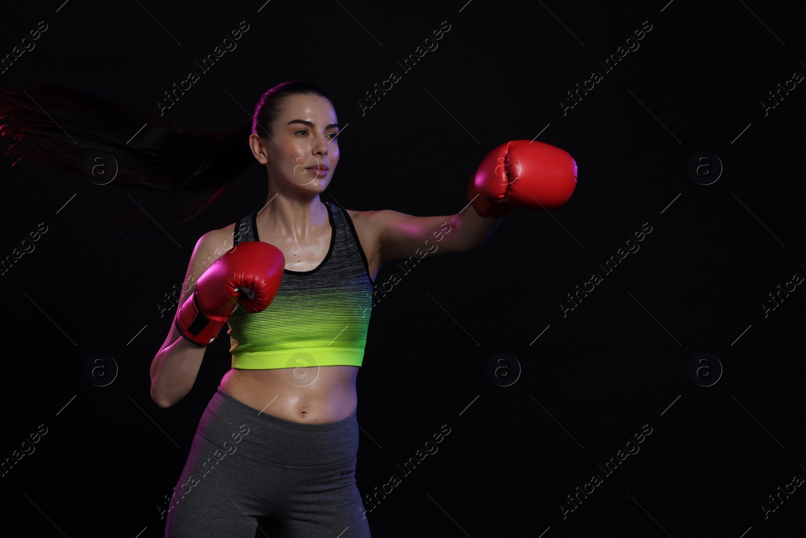 Photo of Beautiful woman wearing boxing gloves training in color lights on black background