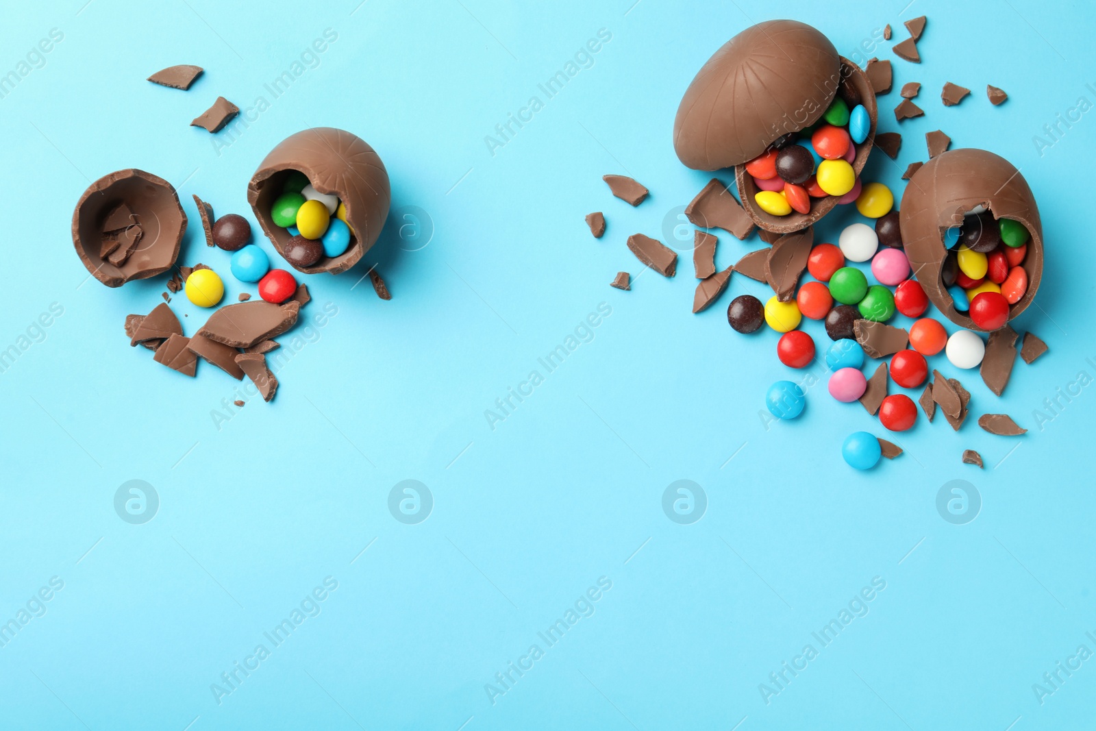 Photo of Broken chocolate eggs with candies on light blue background, flat lay. Space for text