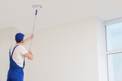 Worker in uniform painting ceiling with roller indoors, low angle view. Space for text