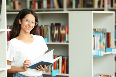 Photo of Young woman with book near shelving unit in library. Space for text