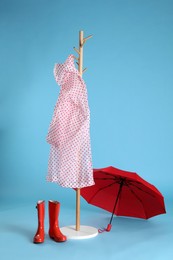 Photo of Open red umbrella, stylish rack with raincoat and rubber boots on light blue background