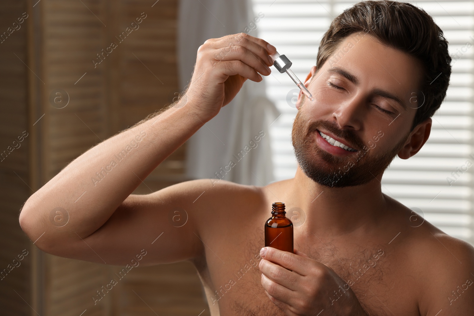 Photo of Smiling man applying cosmetic serum onto his face indoors