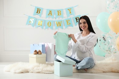 Photo of Happy pregnant woman holding onesie in room decorated for baby shower party