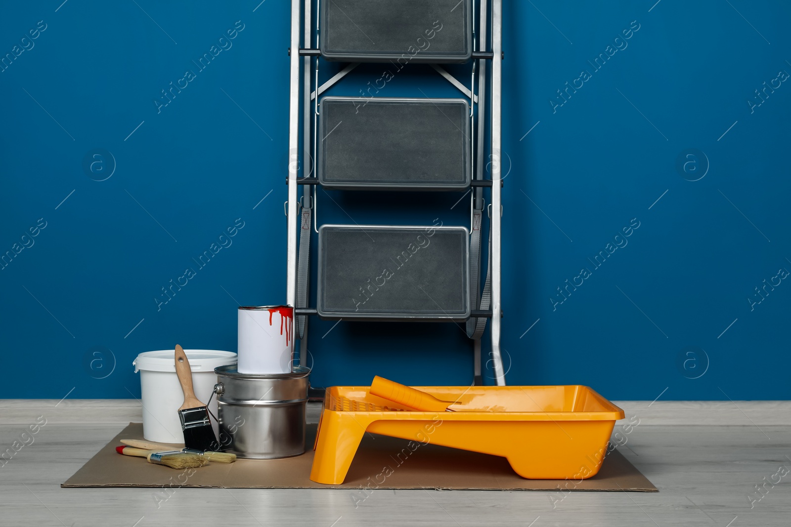 Photo of Metallic folding ladder and painting tools near blue wall indoors
