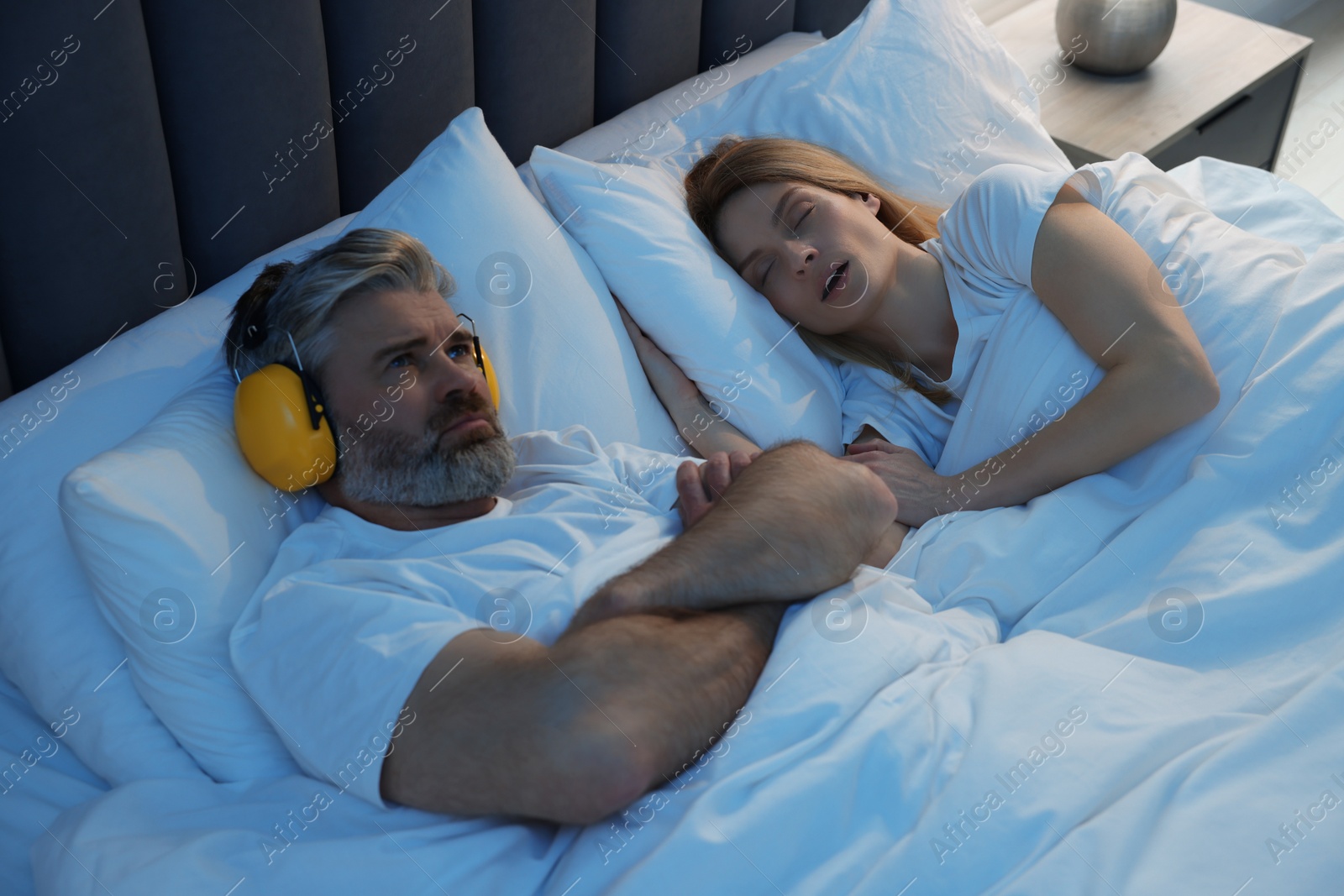 Photo of Irritated man with headphones lying near his snoring wife in bed at home