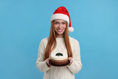 Young woman in Santa hat with tasty cake on light blue background