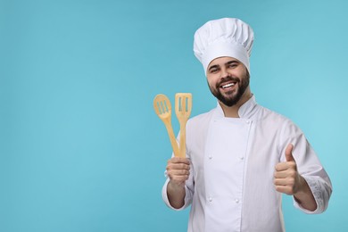 Photo of Happy young chef in uniform holding wooden utensils and showing thumb up on light blue background. Space for text