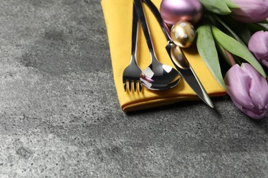 Photo of Cutlery set, painted eggs and beautiful flowers on grey table, closeup with space for text. Easter celebration