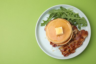 Tasty pancakes with butter, fried bacon and fresh arugula on light green background, top view. Space for text