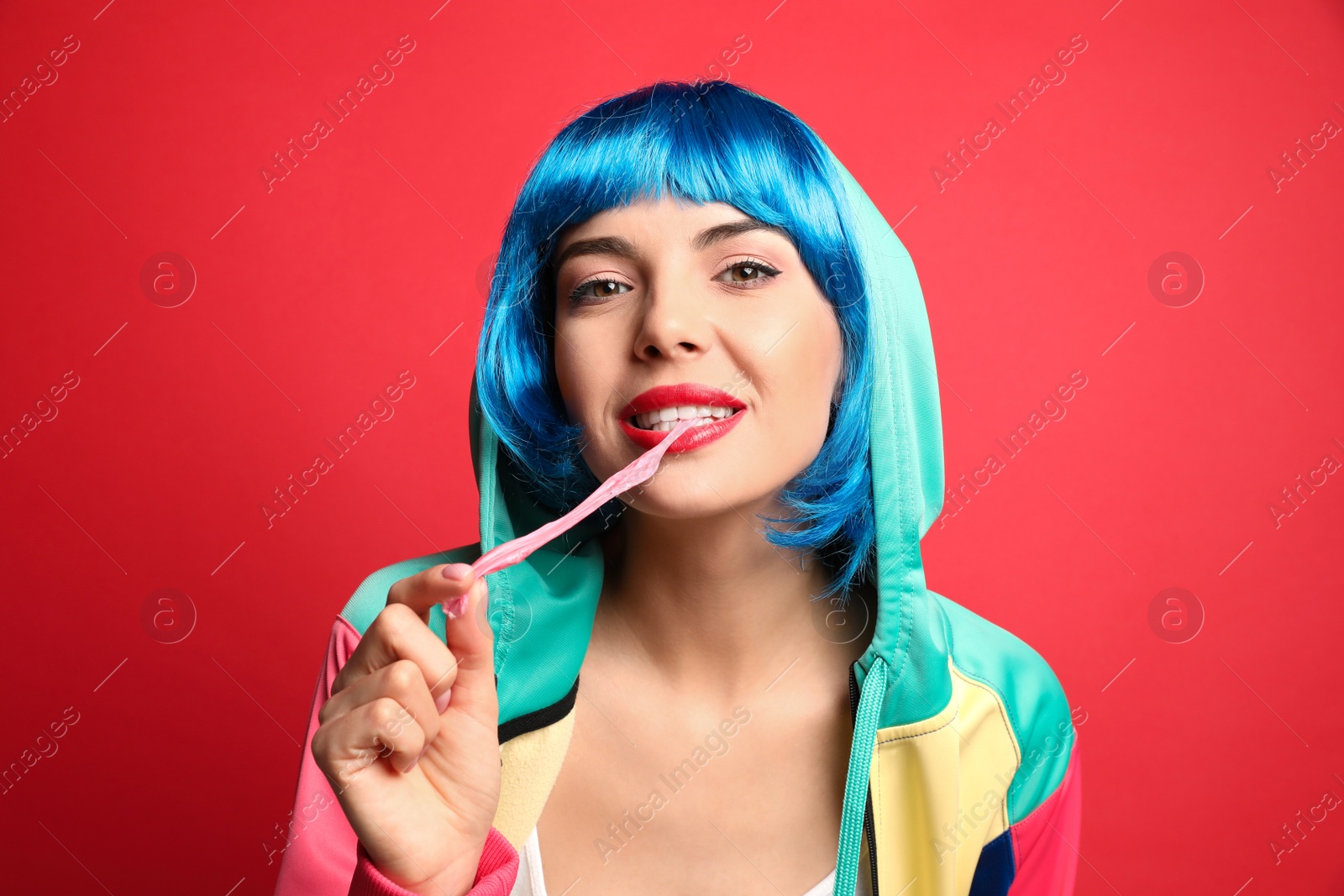 Photo of Fashionable young woman in colorful wig chewing bubblegum on red background