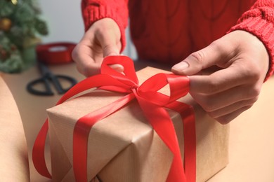 Christmas present. Woman tying ribbon bow on gift box at table, closeup