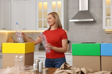 Photo of Garbage sorting. Smiling woman throwing plastic bottle into cardboard box in kitchen