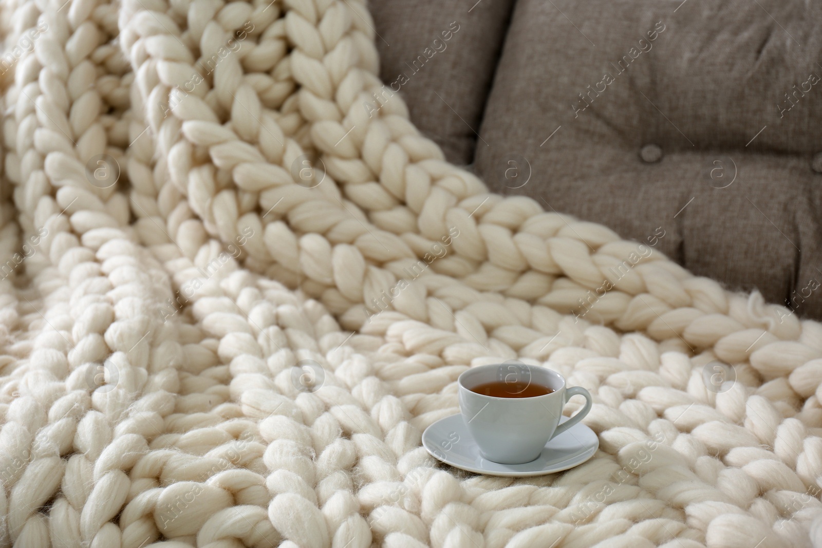 Photo of Cup of drink on couch with soft knitted blanket in room, closeup. Interior element