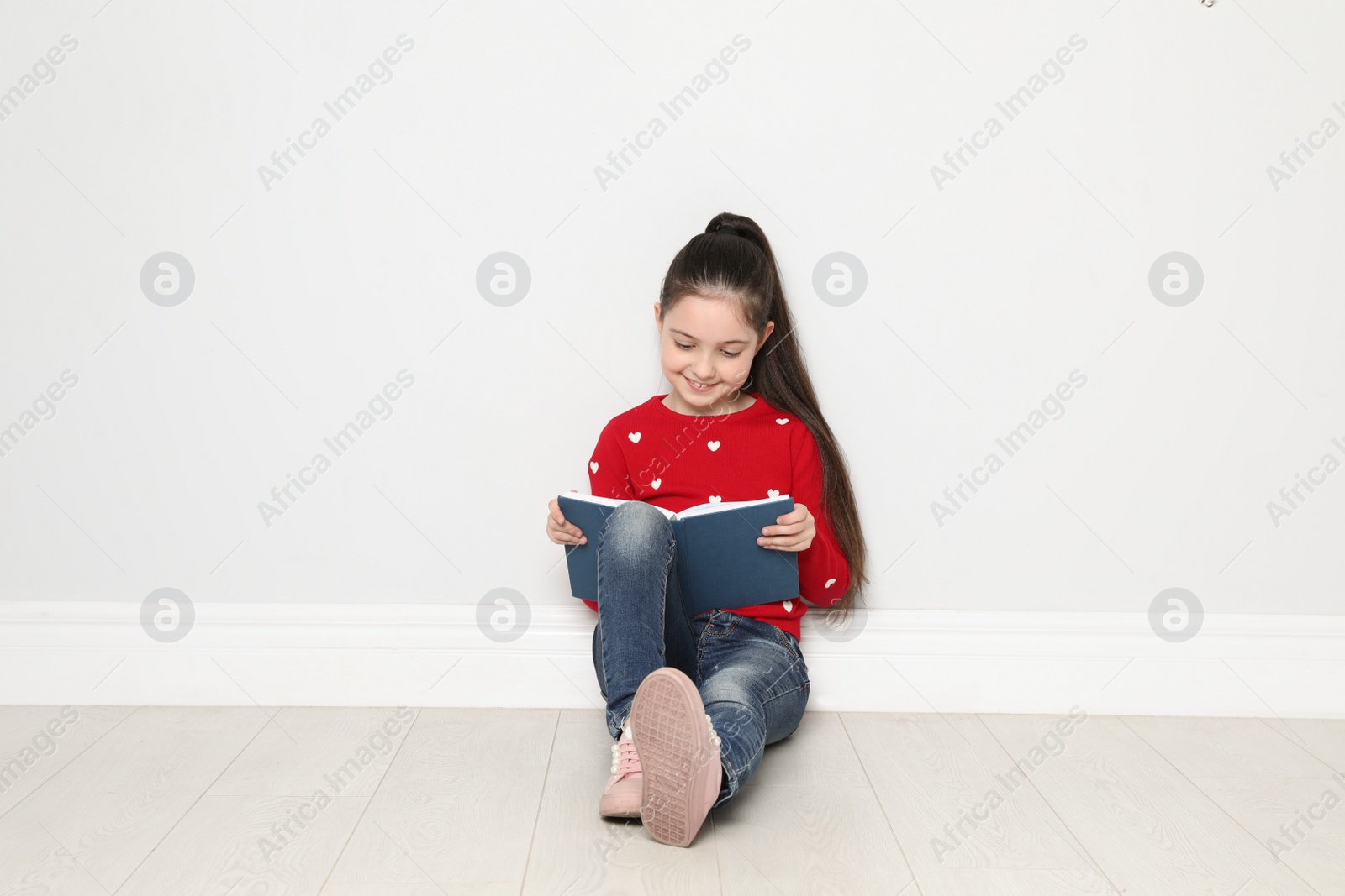Photo of Cute little girl reading book on floor near white wall, space for text