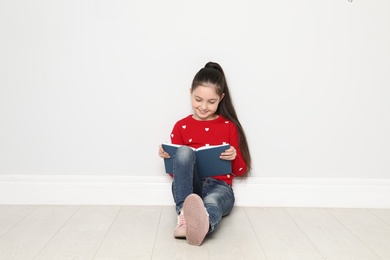 Cute little girl reading book on floor near white wall, space for text