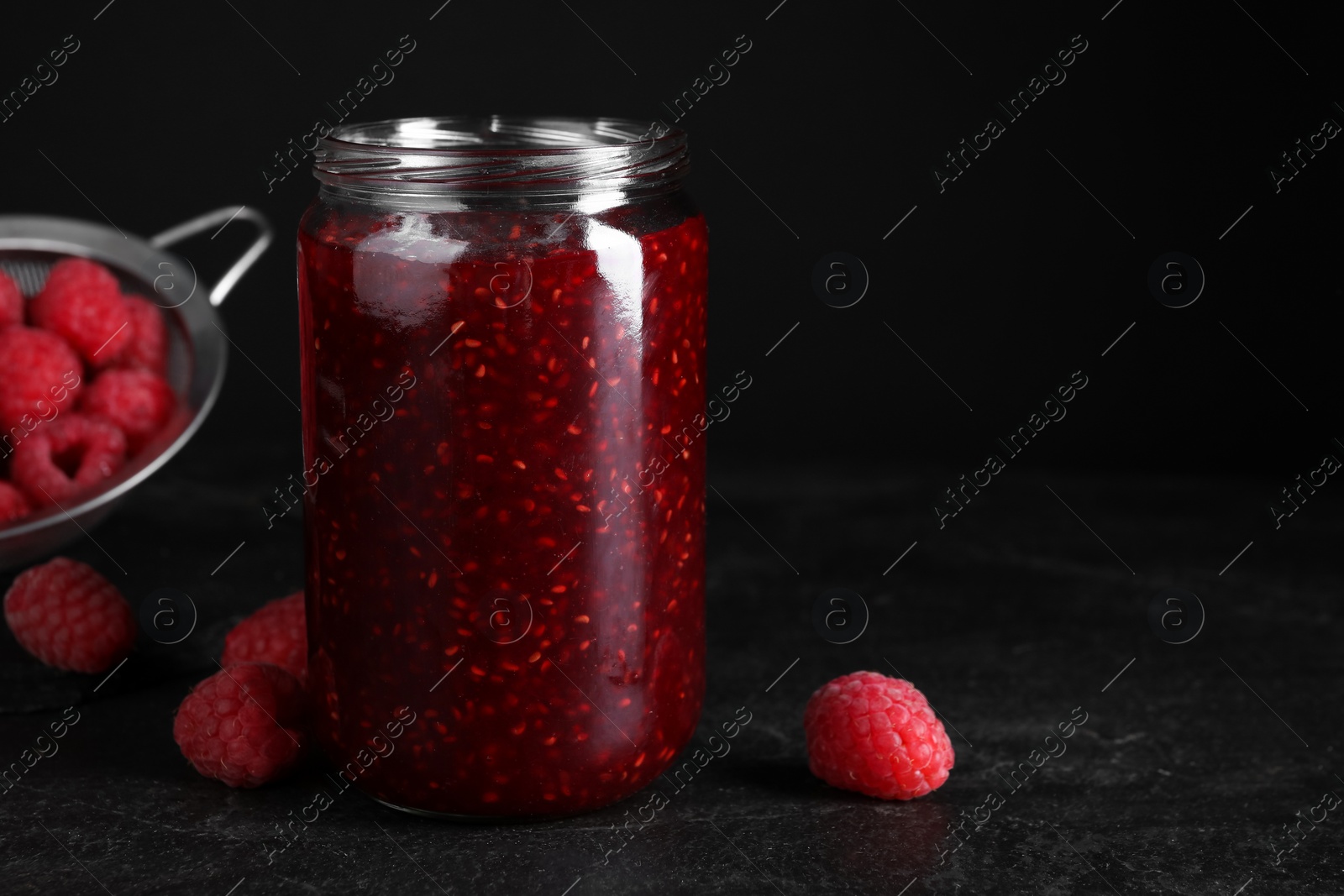 Photo of Delicious jam and fresh raspberries on black slate table, closeup. Space for text